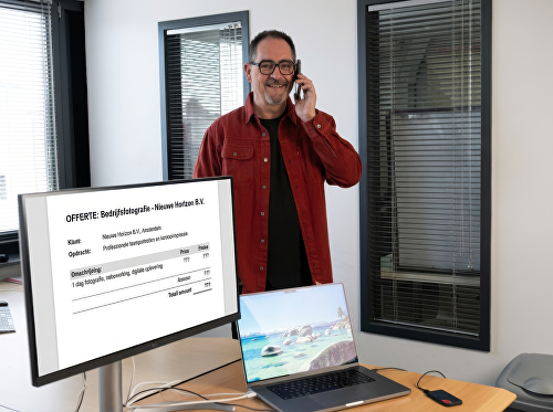 Een fotograaf staat lachend te telefoneren. Op het scherm van de computer een offerte voor Bedrijfsfotografie.